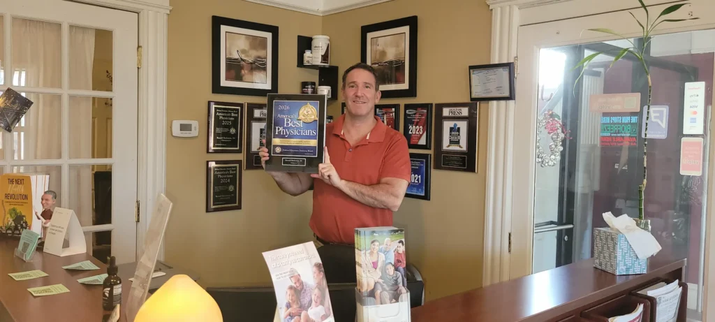 Dr. Russell Brokstein holding up his 2026 America's Best Physicians award standing behind his front desk in Freehold NJ